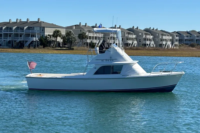 The Image of 1970 Bertram 31 Flybridge Cruiser on calm water, clear sky, residential backdrop. - 0