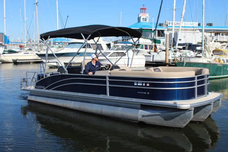 The Image of 2025 Harris Cruiser 230 pontoon boat docked at a marina with a person at the helm. - 0