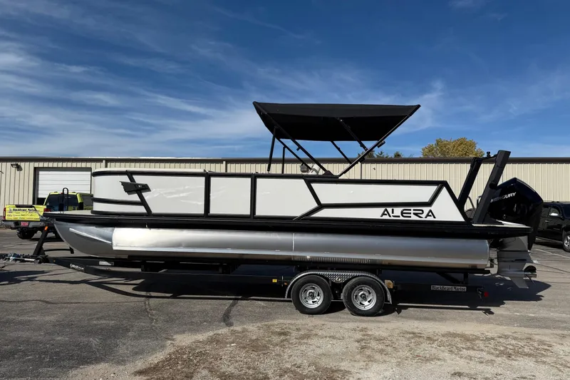 Slide: The Image of 2026 Alera M 25L pontoon boat on trailer, parked outdoors under clear sky. - 2