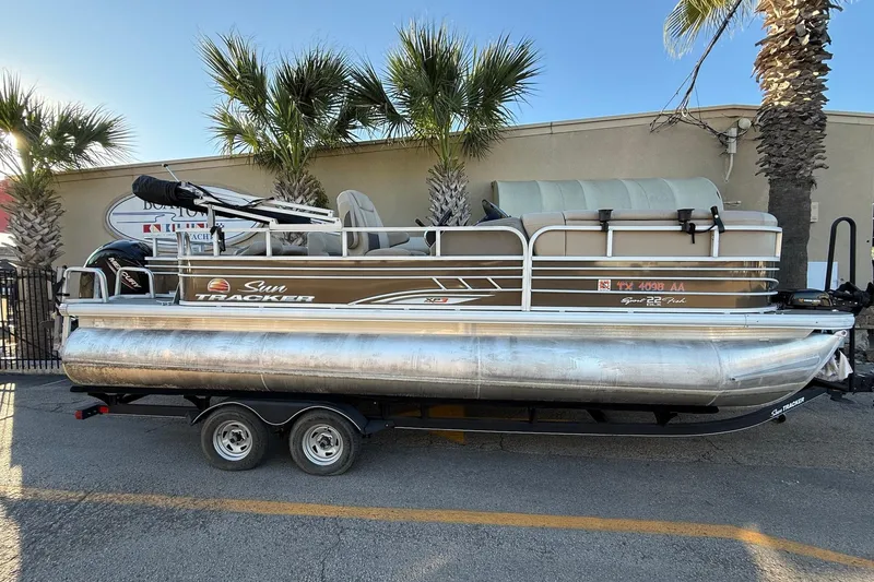 The Image of 2020 Sun Tracker Fishin' Barge 22 XP3 pontoon boat on trailer, parked near palm trees. - 1