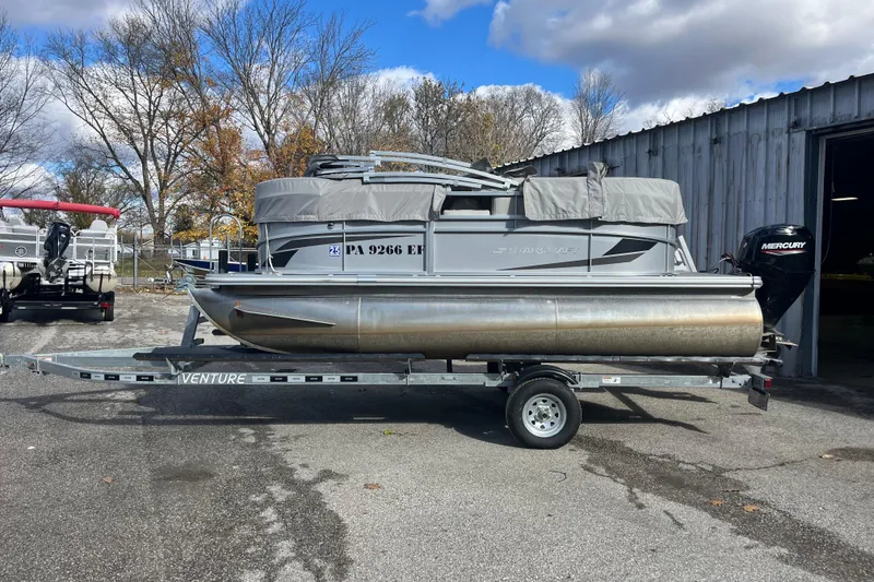Slide: The Image of 2024 Starcraft LX 16 R pontoon boat on trailer, parked outdoors near a metal building. - 1
