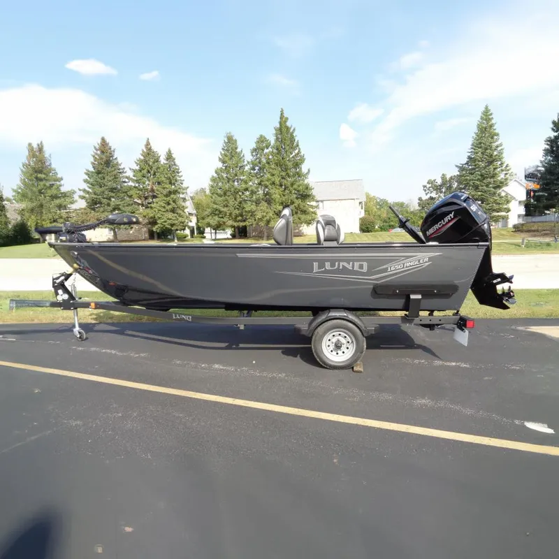 The Image of 2026 Lund 1650 Angler Tiller boat on trailer, parked outdoors, with trees in background. - 0