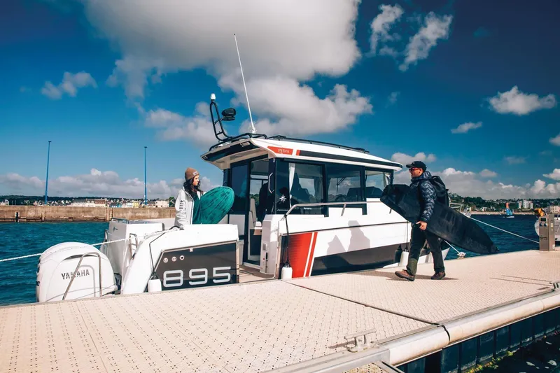 Slide: The Image of 2026 Jeanneau NC895 Sport boat docked, people carrying surfboards, clear sky. - 9