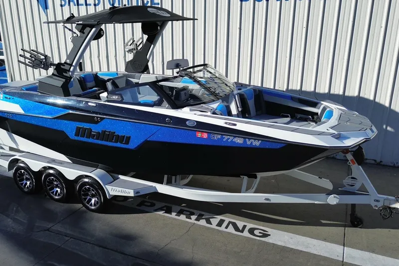 Slide: The Image of 2023 Malibu M240 boat on trailer, blue and black design, parked outdoors. - 2