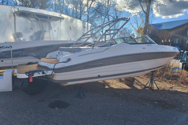 Slide: The Image of 2018 Rinker QX23 BR boat on stands, outdoors, with trees and a building in the background. - 2
