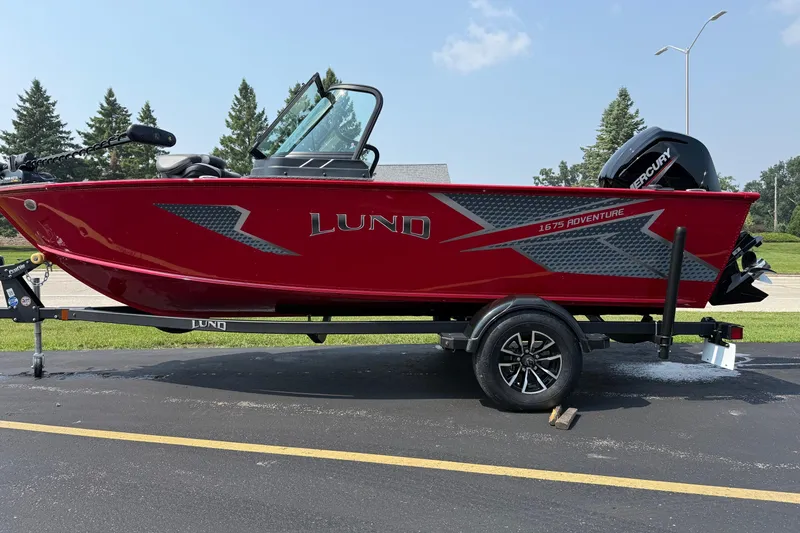 Slide: The Image of 2025 Lund 1675 Adventure Sport boat in vibrant red on a trailer, parked outdoors. - 1