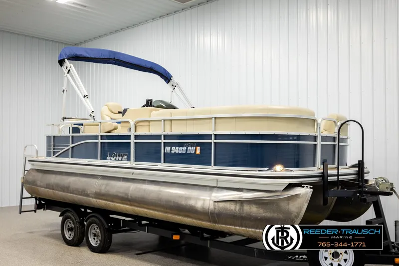 Slide: The Image of 2015 Lowe SS 210 pontoon boat with blue canopy on trailer in indoor storage. - 2