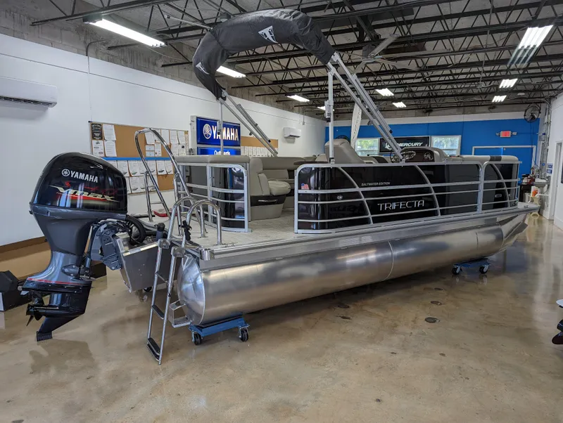 Slide: The Image of 2022 Trifecta 20C LE pontoon boat displayed at a dealership under a sunny sky. - 2