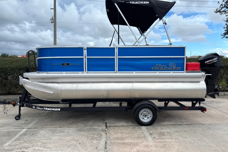 The Image of 2026 Sun Tracker Party Barge 16 DLX pontoon boat on trailer, blue and silver design. - 0