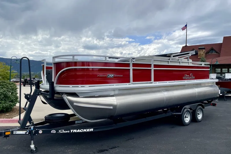 The Image of 2026 Sun Tracker Fishin' Barge 22 XP3 pontoon boat on trailer, red and silver design. - 0