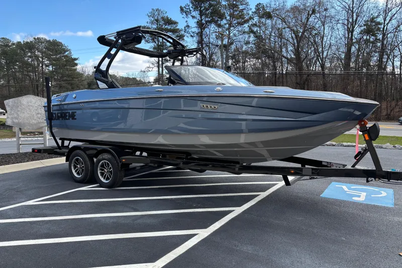 Slide: The Image of 2026 Supreme S220 boat on trailer in parking lot, surrounded by trees. - 2