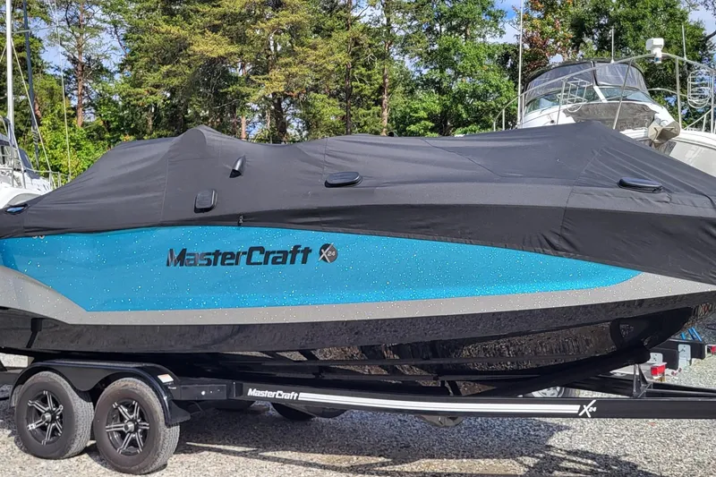 Slide: The Image of 2022 MasterCraft X24 boat covered, on trailer, surrounded by trees and other boats. - 1