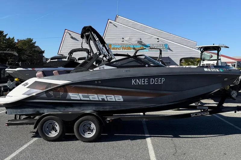 The Image of 2017 Scarab 215 HO Impulse boat on trailer, parked outdoors, with "Knee Deep" text on side. - 1