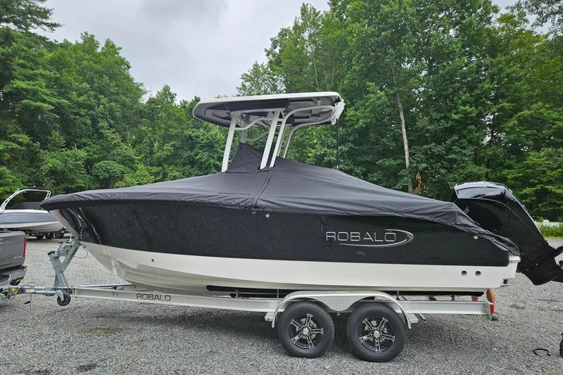 Slide: The Image of 2025 Robalo R230 Center Console boat covered, on trailer, surrounded by trees. - 2