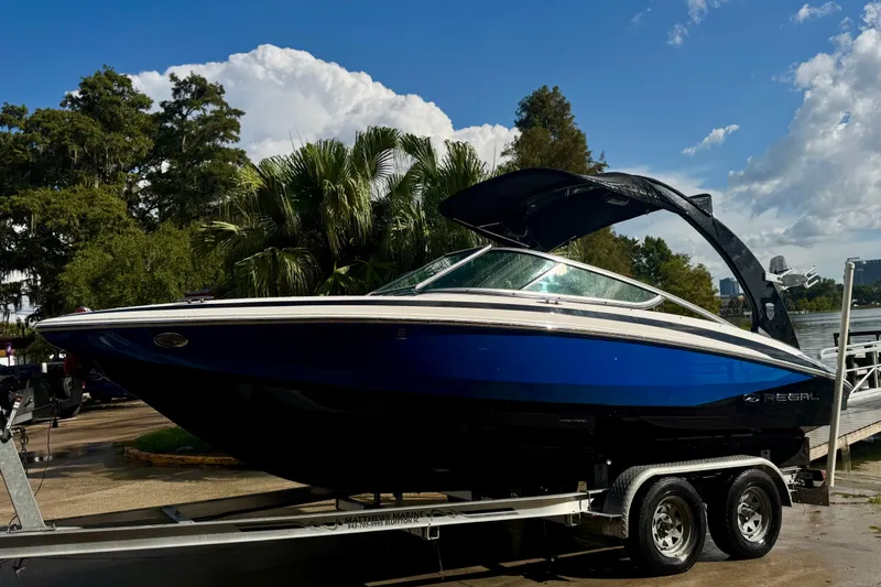 The Image of 2013 Regal 21 OBX boat on trailer, parked near lush greenery and water. - 1