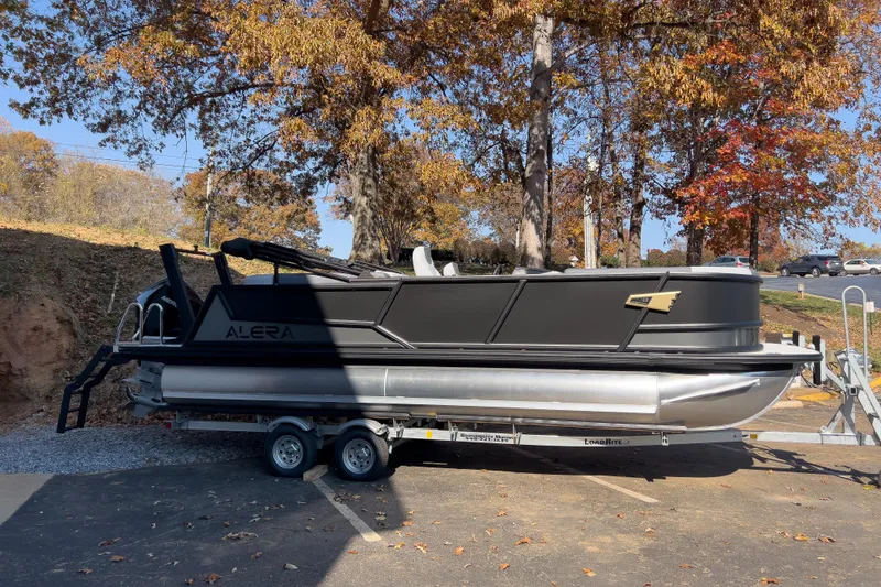 Slide: The Image of 2026 Alera M23Q pontoon boat on trailer, parked under autumn trees. - 2