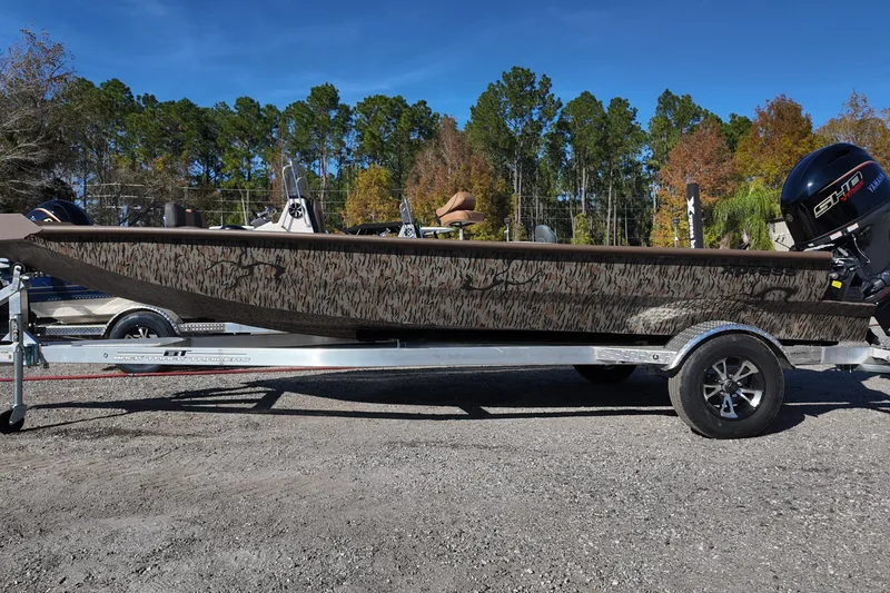 Slide: The Image of 2026 Xpress X18DB boat on trailer, camo design, parked outdoors with trees in background. - 2