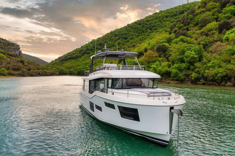 Slide: The Image of 2025 Beneteau Grand Trawler 63 cruising on a scenic river with lush green hills. - 22