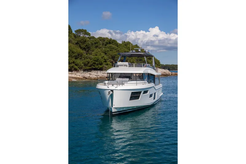 Slide: The Image of 2025 Beneteau Grand Trawler 63 yacht on serene blue water near a lush coastline. - 23