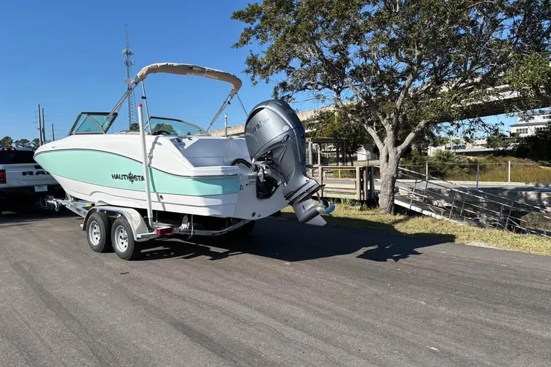 Slide: The Image of 2026 NauticStar 223 DC Deck boat on trailer, parked near a tree and waterway. - 3
