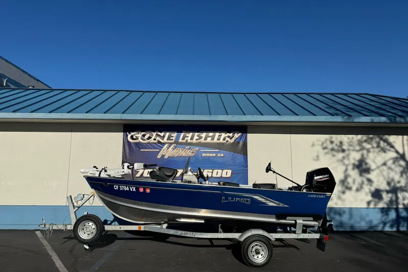 Slide: The Image of 2024 Lund 1400 Fury SS boat on trailer, parked outside a marine store. - 0