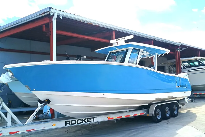 The Image of 2026 Robalo R360 Center Console boat on trailer, blue hull, parked under shelter. - 0
