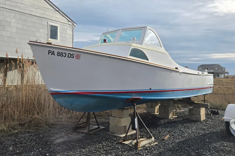 The Image of 1964 Whiticar 24 boat on blocks, white hull with blue and red trim, near coastal house. - 0
