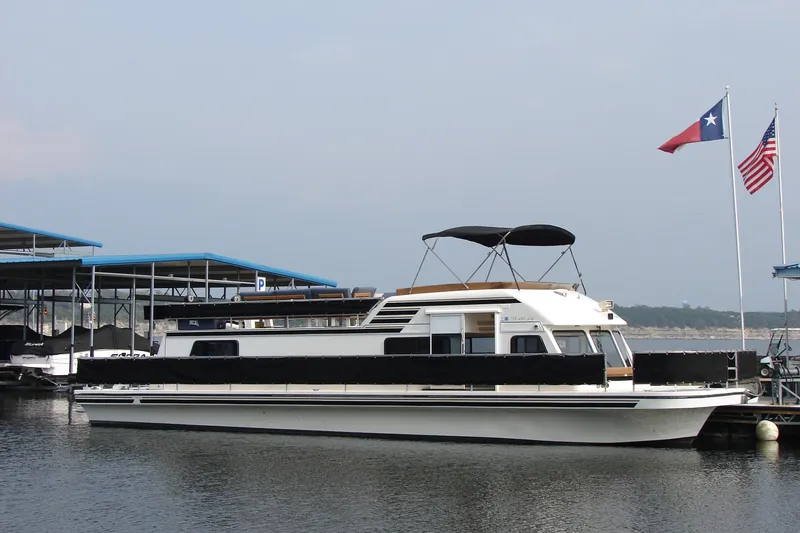 Slide: The Image of 1997 Gibson 47 Sport Houseboat docked with flags, featuring a covered upper deck. - 47