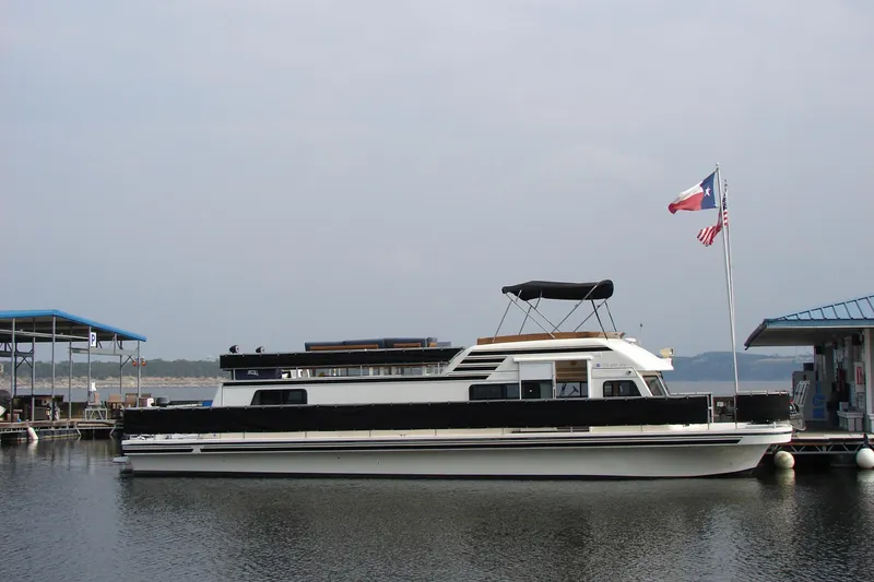 Slide: The Image of 1997 Gibson 47 Sport Houseboat docked with flags, calm water, and overcast sky. - 48