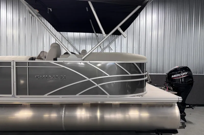 Slide: The Image of 2026 Sylvan Mirage 8520 LZ pontoon boat with Mercury outboard motor in a metal-walled showroom. - 21