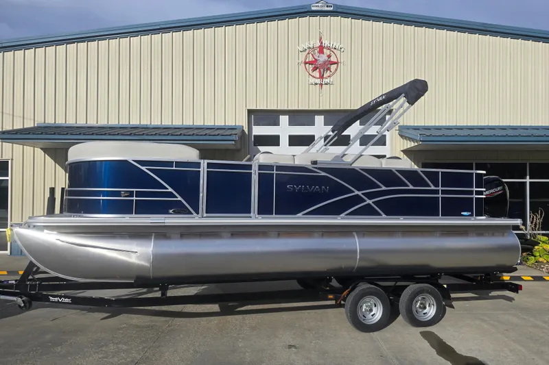 Slide: The Image of 2025 Sylvan Mirage Fish 8522 Party Fish pontoon boat on trailer, parked outside a building. - 1