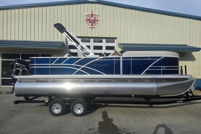 Slide: The Image of 2025 Sylvan Mirage Fish 8522 Party Fish pontoon boat on trailer, parked outside a marine building. - 8