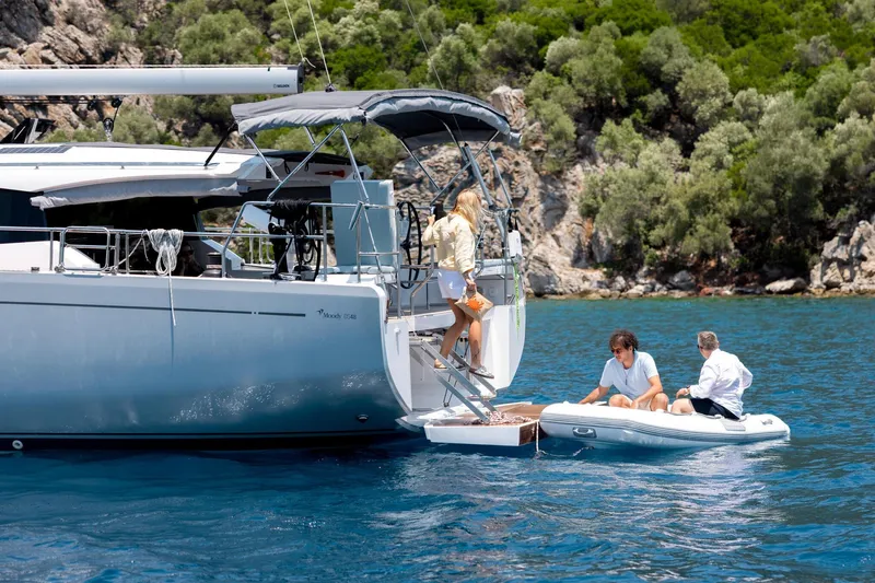 Slide: The Image of 2026 Moody 48 Deck Saloon yacht with people boarding from a small boat, scenic backdrop. - 18