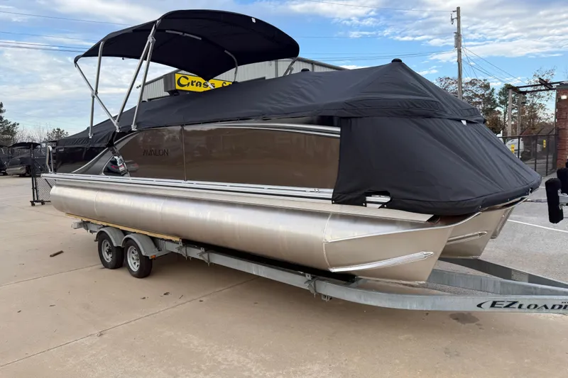 Slide: The Image of 2026 Avalon LSZ Versatile Rear Bench pontoon boat on trailer, covered and parked outdoors. - 6