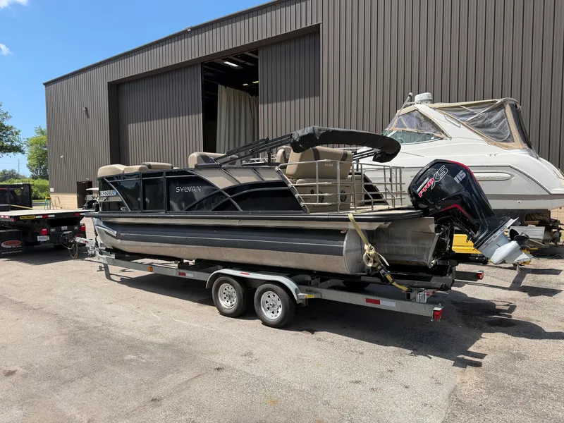 Slide: The Image of A 2022 Sylvan L1 DLZTT pontoon boat on a trailer in front of a large building. - 1