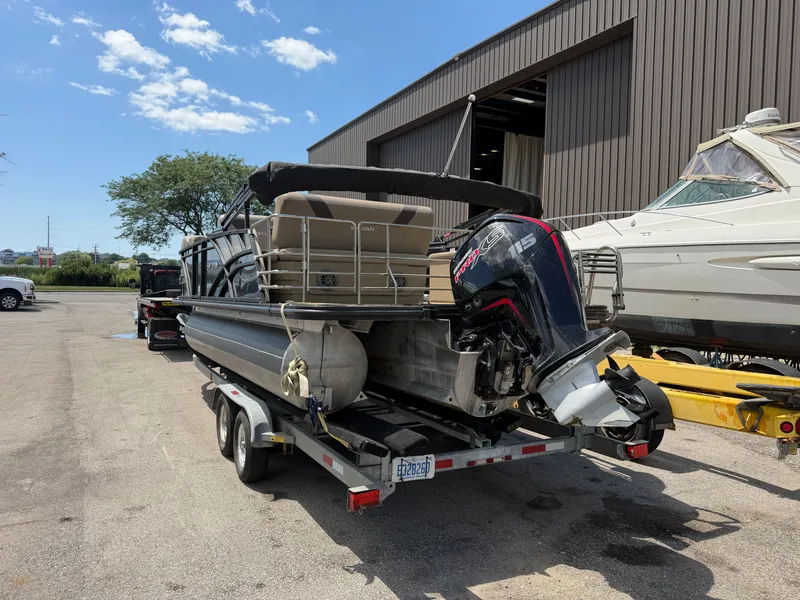 Slide: The Image of A 2022 Sylvan L1 DLZTT pontoon boat on a trailer, parked beside a building. - 2