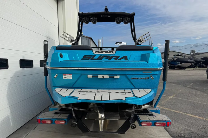 Slide: The Image of 2021 Supra SA 575 boat, blue, parked outside a garage, rear view. - 2