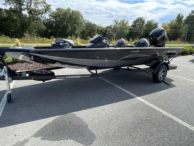 Slide: The Image of 2023 Vexus AVX181 boat on trailer in parking lot, surrounded by trees. - 2