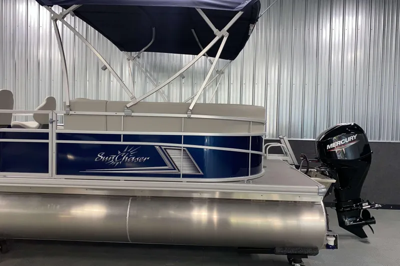 Slide: The Image of 2026 SunChaser Vista 18 Fish pontoon boat with Mercury outboard motor in a metal-walled showroom. - 19
