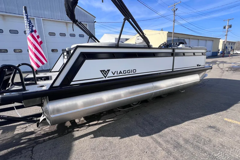Slide: The Image of 2026 Viaggio V 20U pontoon boat with American flag, parked near industrial buildings. - 2