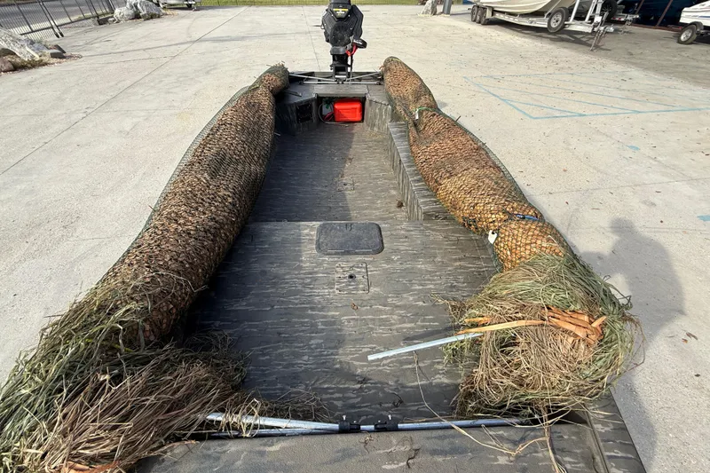 Slide: The Image of War Eagle 750 Gladiator 2026 boat with camouflage netting and grass, parked on concrete. - 9