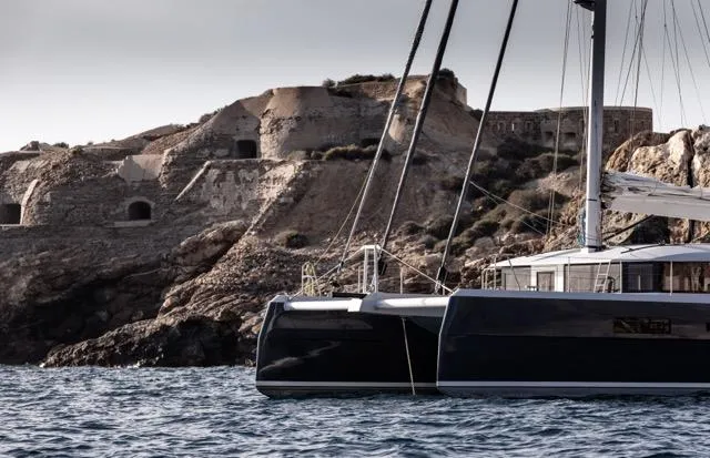 Slide: The Image of Catamaran sailing near rocky coastline, Eyecat 55 Autumn 2024 model, under a cloudy sky. - 7