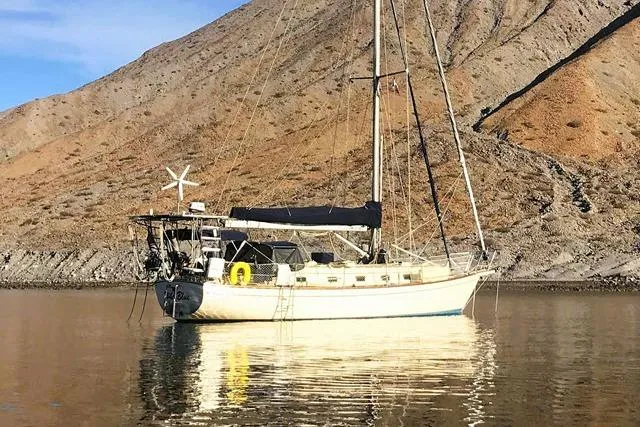 The Image of 1999 Island Packet 45 sailboat anchored near rocky shoreline under clear blue sky. - 0