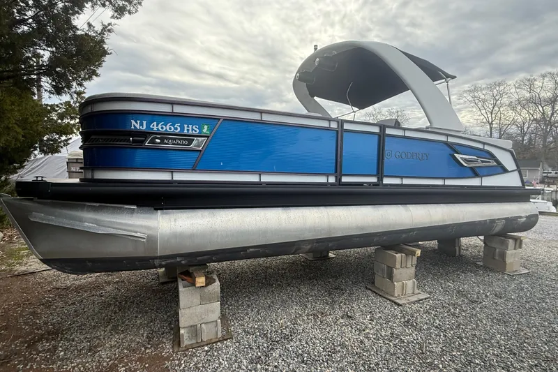 The Image of 2023 Godfrey AquaPatio 255 ULC pontoon boat on blocks, blue exterior, overcast sky. - 0
