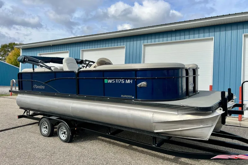 The Image of 2018 Manitou 23 Aurora LE pontoon boat on trailer, parked near blue building. - 1