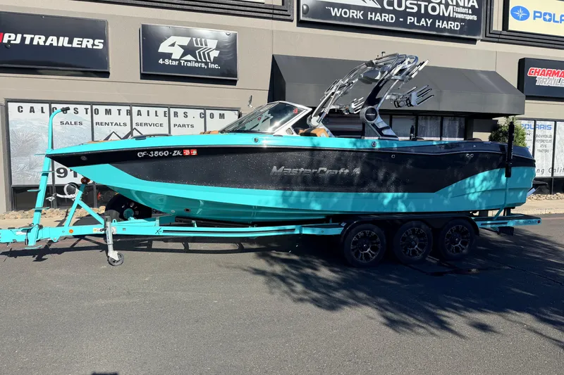 The Image of 2021 MasterCraft X26 boat on trailer outside dealership, vibrant turquoise and black design. - 0