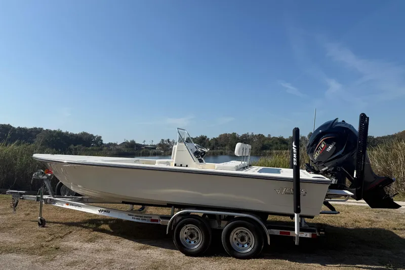 Slide: The Image of 2026 Hewes 21 REDFISHER boat on trailer, parked near a lake under clear blue sky. - 2