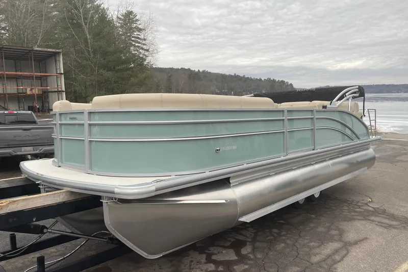 Slide: The Image of 2026 Harris Cruiser 230 pontoon boat on trailer, parked near a lake with overcast skies. - 2