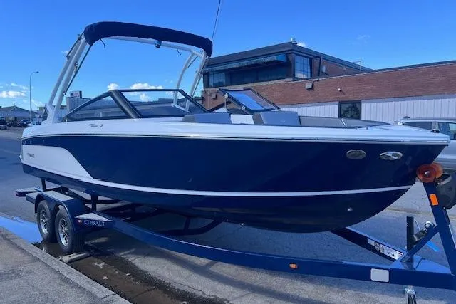 Slide: The Image of 2025 Cobalt CS23 Surf boat on trailer, parked outdoors, blue and white design. - 1