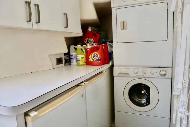 Slide: The Image of Laundry area with stacked washer-dryer, cleaning supplies, and white cabinets on a Westport 112 yacht. - 49
